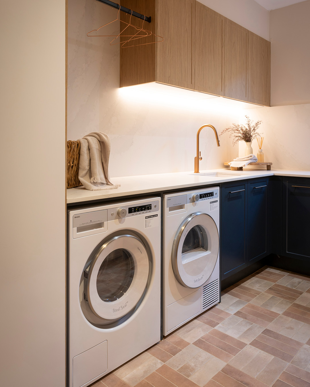 Laundry with oak and navy blue cabinetry, hnaging rail, laundry appliances, and sink and tap.
