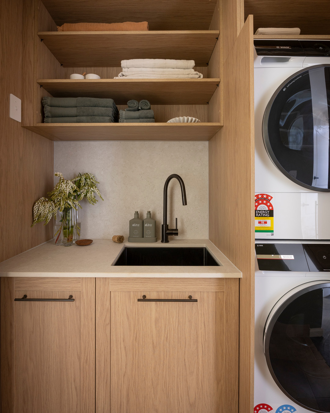 Small laundry with stacked washing machine and clothes dryer, storage space and sink and tap.